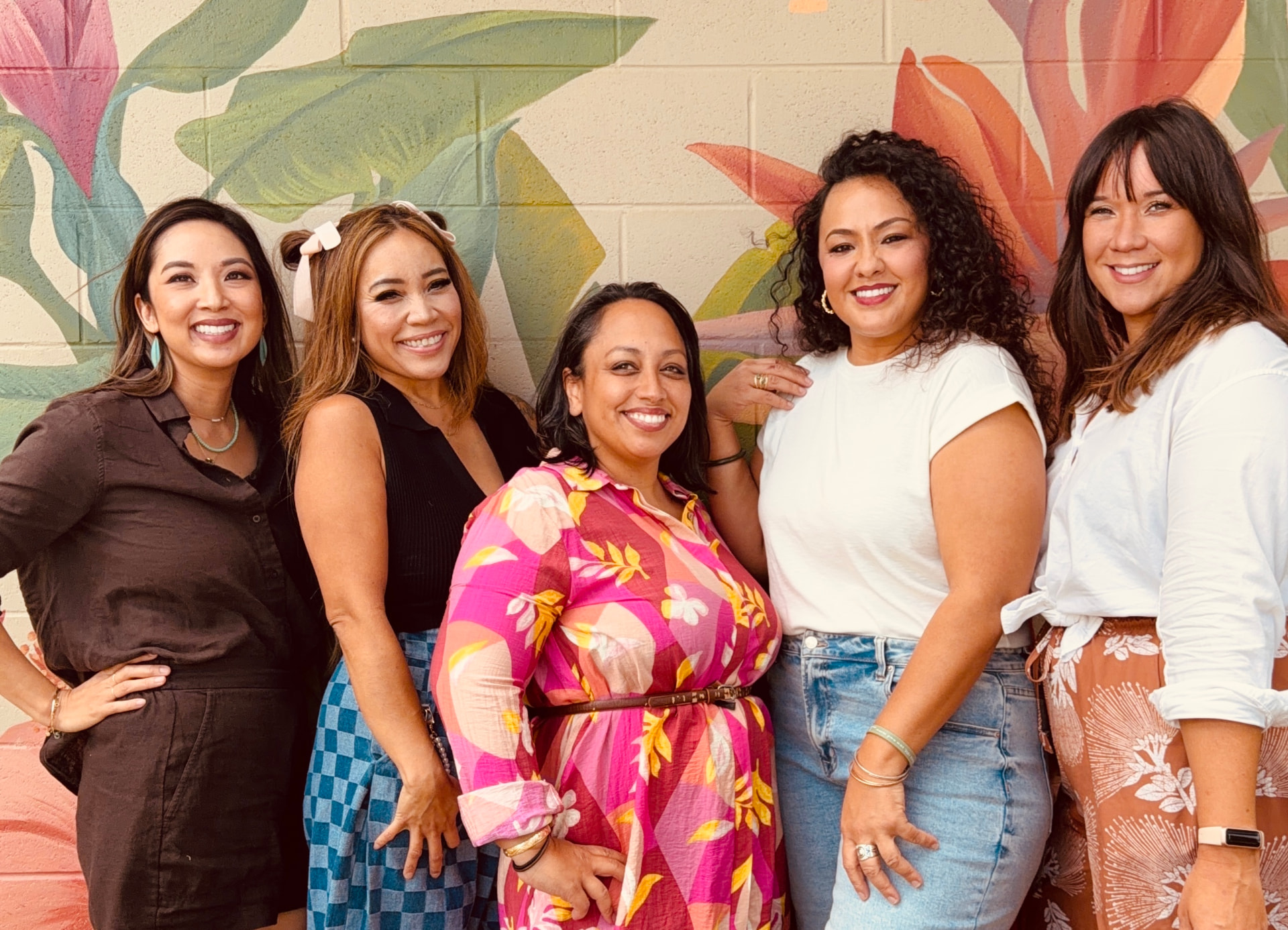 Five women smiling together in front of a painted tropical mural in Honolulu